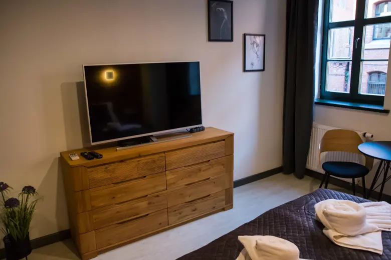 Salon z telewizorem i drewnianą komodą, przy oknie stoi stolik z krzesłami.