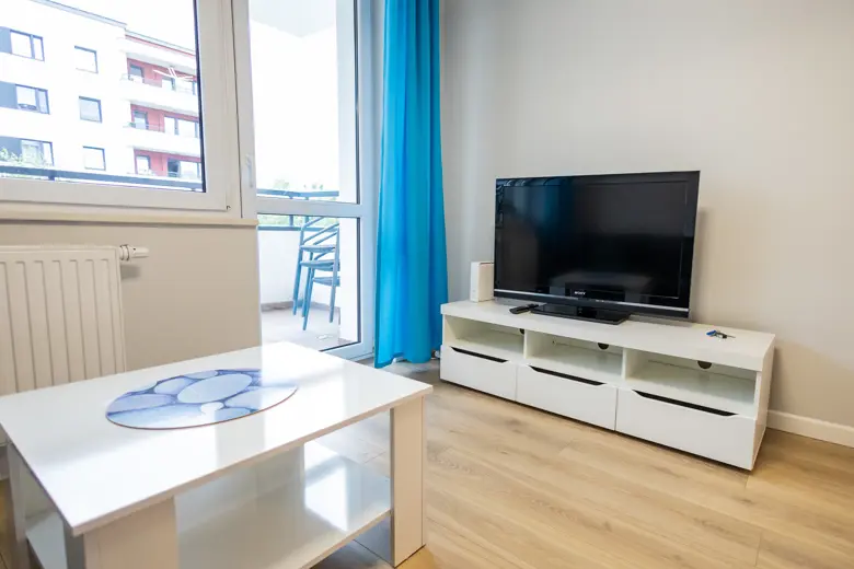 Salon z telewizorem na białej szafce i jasnym balkonowym światłem.