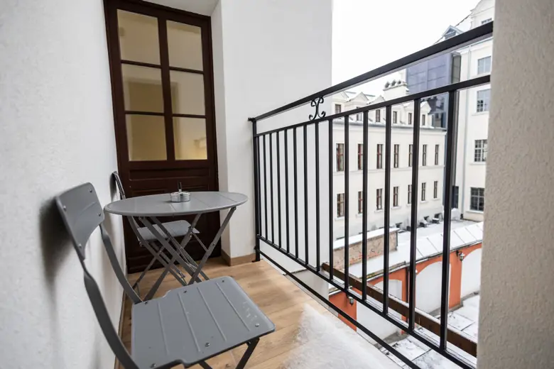 Balkon z metalowym poręczem, mały stół i dwa krzesła, widok na zewnętrzny gzyms budynków.