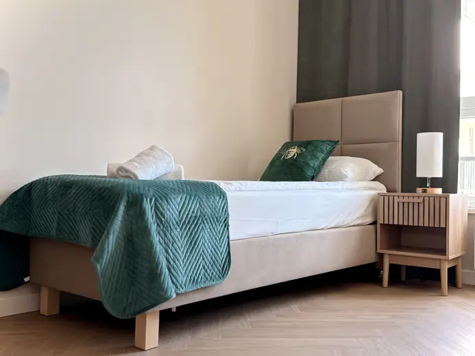Widok na bok łóżka z zieloną narzutą i lampą nocną.
