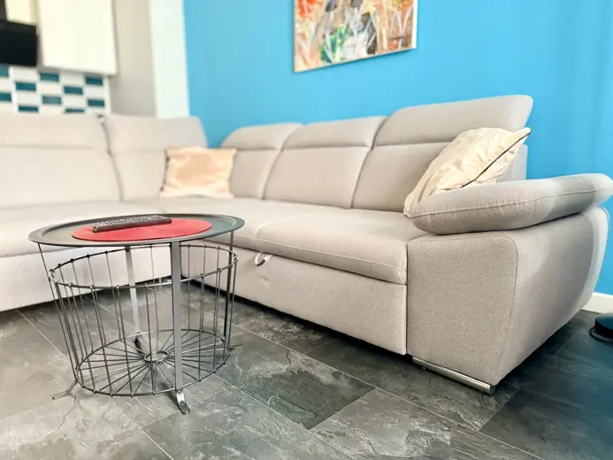Sofa narożna w jasnym beżu, przy błękitnej ścianie.