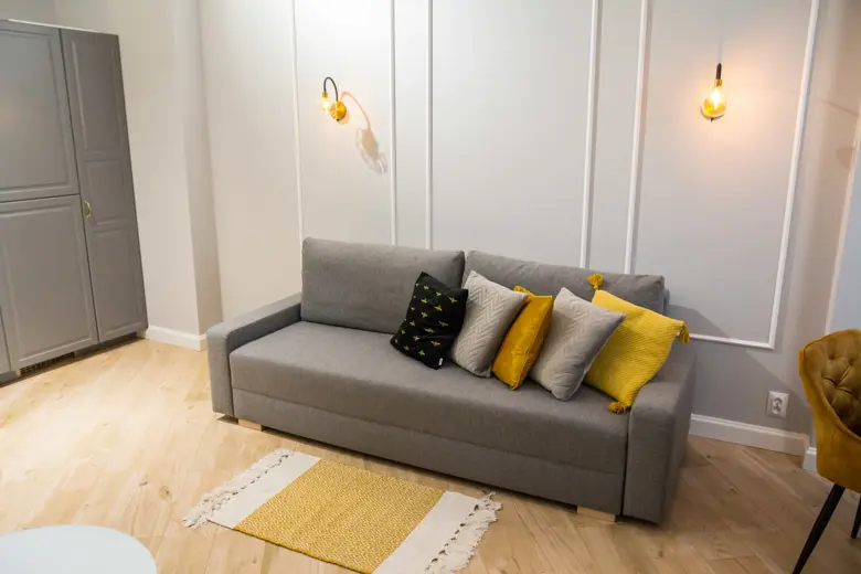 Szara sofa z żółtymi poduszkami, białe panele i lampki.