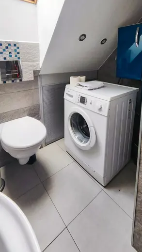 Pralka przy ścianie obok toalety w łazience, biała ceramika na tle jasnych płytek.