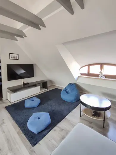 Salon z belkami sufitowymi, telewizorem na białej szafce i niebieskimi pufami