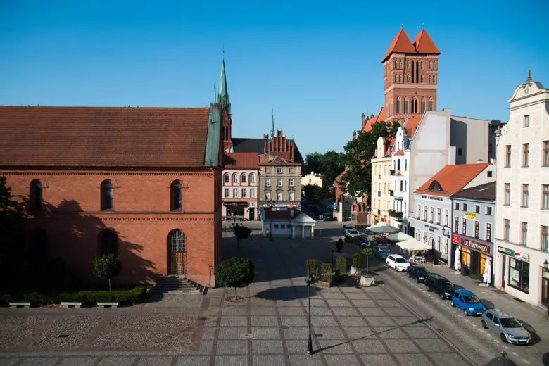 Uliczny widok z kamienic i czerwonych dachów oraz chodnikiem na planie kwadratu.