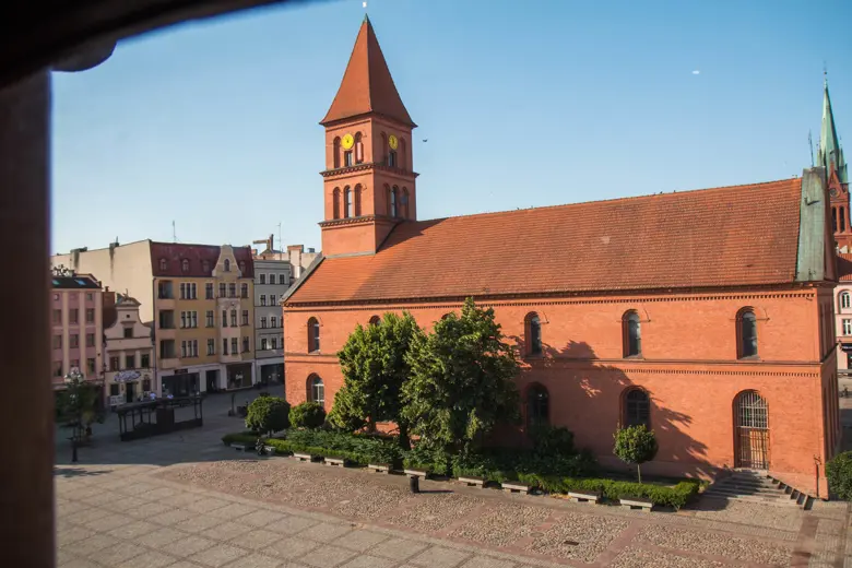 Kościół z czerwonej cegły na placu, otwarte alejki i zabudowa wokół.