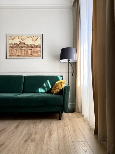 Zielona sofa z żółtą poduszką, lampą stojącą obok i jasnymi zasłonami.